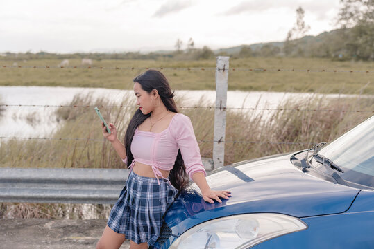 A beautiful lady is standing on the side of the road leaning on the hood of a blue sedan car, holding her phone, checking and scrolling to call someone for help.