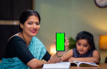 Happy smiling mother showing green screen mobile phone by looking camera in front of reading kid at home - concept of app promotion, advertisement and online education