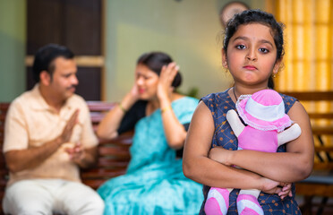 Rack focus shot of sad kid with doll in front shouting and arguing parents on sofa at home - concept of depression, innocence and relationshiop problems