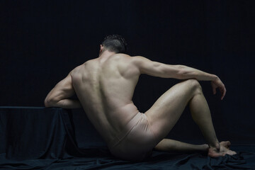Statue. Back view portrait of muscular shirtless man sitting and posing in underwear against black...