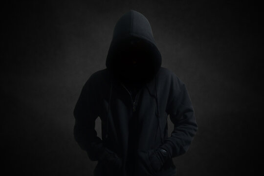 Mysterious man wearing black hoodie standing against dark background. Hacker, crime, and cyber security concept.