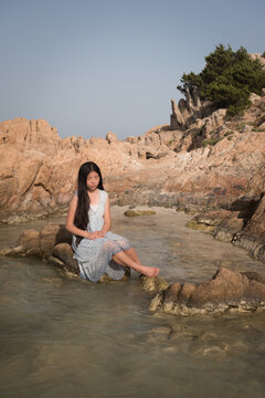 fine art portrait of asian woman in a dress sitting on rock in ocean