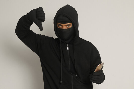 Portrait of mysterious man wearing black hoodie and mask doing hacking activity on mobile phone, hacker holding a smartphone. Isolated image on gray background