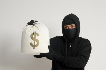 Mysterious robber thief man wearing black hoodie and mask carrying a bag full of money. Isolated image on gray background