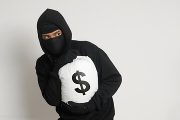 Mysterious robber thief man wearing black hoodie and mask carrying a bag full of money. Isolated...