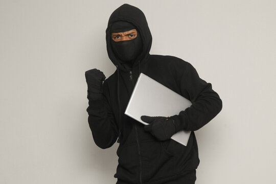 Mysterious robber thief man wearing black hoodie and mask stealing laptop while raising his fist, celebrating success. Isolated image on gray background