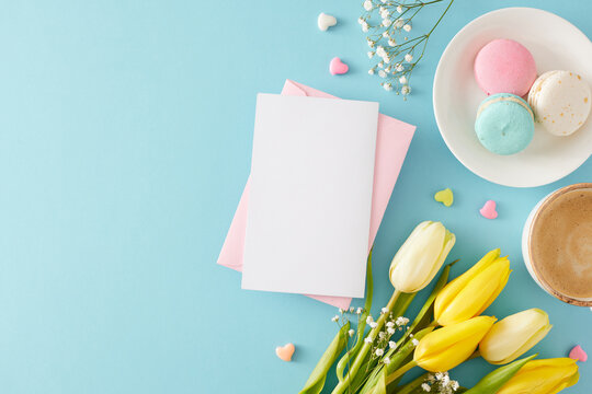 Women's Day atmosphere concept. Top view photo of envelope with postcard macaroons cup of coffee small hearts yellow white tulips and gypsophila flowers on pastel blue background with copyspace