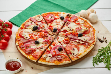Freshly baked pizza with smoked sausages, red pepper, mushrooms, onion and olives served on wooden background with tomatoes, sauce and herbs. Food delivery concept. Restaurant menu