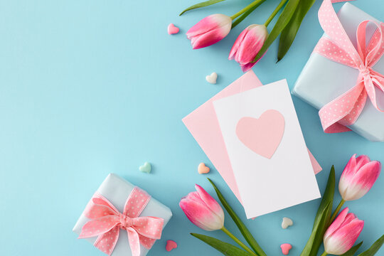 Mother's Day celebration concept. Flat lay photo of postcard with heart gift boxes small hearts baubles and pink tulips on isolated pastel blue background with copy space