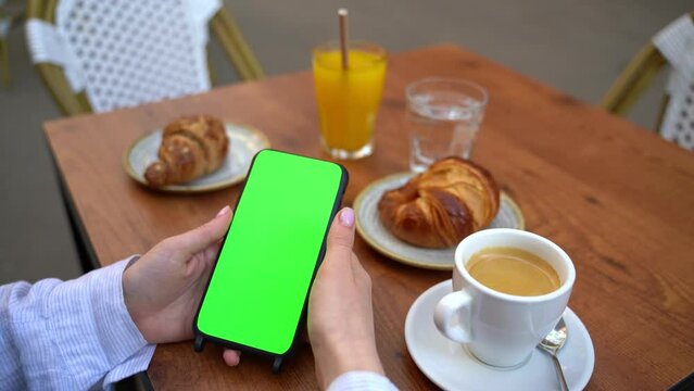chromakey green screen hand holding mobile phone vertical portrait position. Hand swiping left and right. Street cafe breakfast with coffee  and croissant. Template video footage empty copyspace