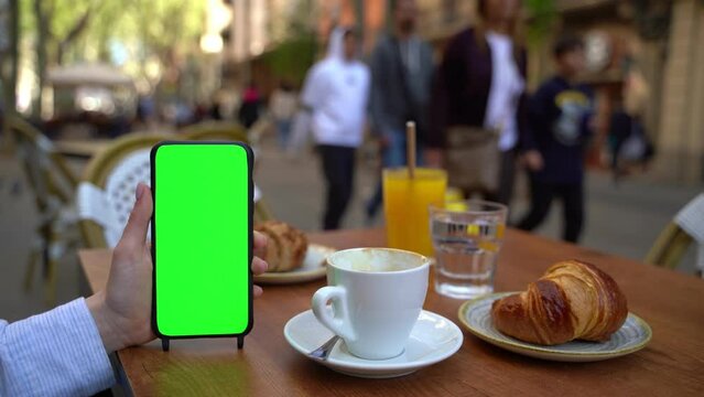 Woman holding cell phone without touching screen during having breakfast in street cafe with coffee and croissant. . Green chroma key screen display. Template moke up video footage