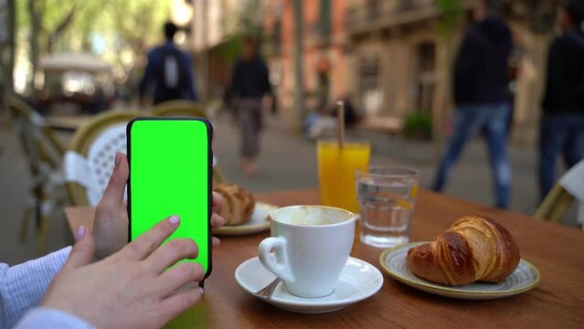 Woman using cell phone during having breakfast in street cafe with coffee and croissant. Scrolling touching the screen. Green chroma key screen display. Template moke up video footage