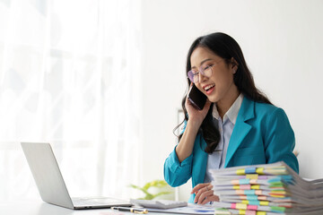 Asian Woman entrepreneur busy with her work in the office. Young Asian woman talking over smartphone or cellphone while working on computer at her desk.