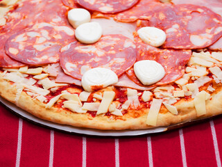 Thin pizza portion with high meat content. Uncooked product with cheese, salami, ham and tomato sous on simple grey red and white stripe table cloth. Dinner preparation. Italian style meal.