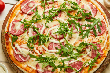 Freshly baked tasty pepperoni pizza with salami, mozzarella cheese and rukkola served on wooden background with tomatoes, sauce and herbs. Food delivery concept. Restaurant menu