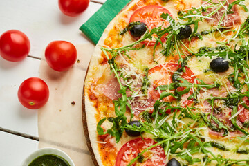 Freshly baked pizza with ham, rukkola, sauce pesto and olives served on wooden background with tomatoes, sauces and herbs. Food delivery concept. Restaurant menu