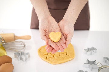 Obraz na płótnie Canvas Woman are cooking holiday cookies in the kitchen, lifestyle photo series, hands closeup only. Female hands holding dough in heart shape. Baking ingredients on the white table in home kitchen.