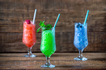 Summer bright desserts of frozen blueberry juice, mint, cucumber and raspberry juice in glass glasses with a straw on a wooden background. Summer cool, tonic crushed ice cocktail, a kind of sherbet