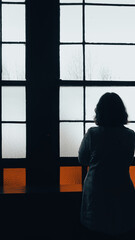 A young woman dressed in  retro-style trench coat stands on the windowsill with her back turned and looks out the window.