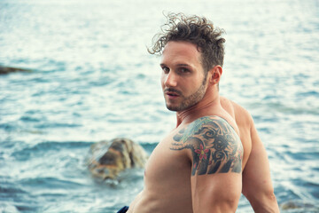 Attractive muscular man sitting by sea or ocean shore, looking at camera