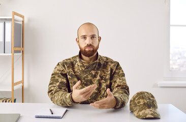 Fototapeta premium Portrait of young soldier during video call. Shaven headed bearded man in uniform sitting at desk in military headquarters office, looking at camera with serious face expression, talking and gesturing