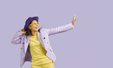 Side view of young stylish modern woman with dark hiar wearing in hat, purple jacket and yellow trousers with glasses waving hand . Half-length portrait isolated on purple background. 