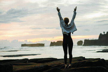 Fototapeta premium A young girl admires the sunset on the ocean, enjoys life, arms outstretched to the sides, rear view.