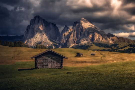 The last rays of sunlight at sunset in Alpe di Siusi. View with small wooden log cabin on green grassland and Langkofel (Sassolungo) mountain range on background. Dolomites, Alto Adige, Bolzano, Italy