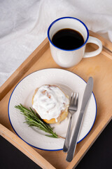 cinnamon roll with rosemary on a white plate, a cup of coffee and cream on a wooden tray