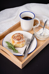 cinnamon roll with rosemary on a white plate, a cup of coffee and cream on a wooden tray