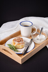 cinnamon roll with rosemary on a white plate, a cup of coffee and cream on a wooden tray