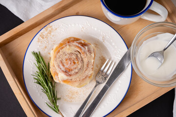 cinnamon roll with rosemary on a white plate, a cup of coffee and cream on a wooden tray