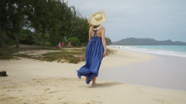 Slow motion scenic shot elegant traveller with lei flower neckless walking in scenic Oahu nature. 30s authentic traveler woman in purple maxi boho style dress and straw hat enjoying Hawaiian beach 4K