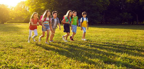 Happy children walking on green park lawn, enjoying free time and good sunny summer weather. Group of cheerful classmates with rucksacks playing, having fun and exploring nature on school field trip