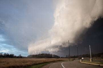 An immense shelf cloud and severe thunderstorm approaches rapidly with a road and streetlights in the foreground.