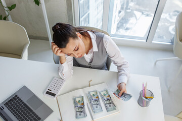 Sad business woman sits at work desk in front of laptop and counts cash. Concept of low salary