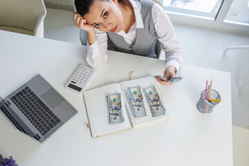 Sad business woman sits at work desk in front of laptop and counts cash. Concept of low salary