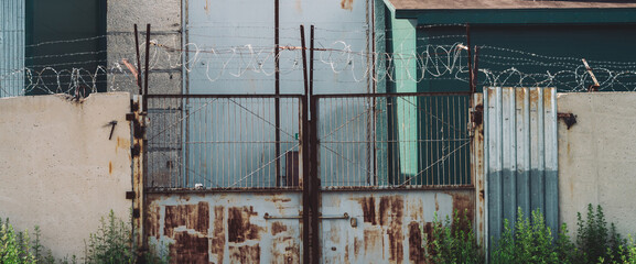 Picturesque industrial building close-up. Metallic rusty gate and concrete fence with barbed wire. Entrance in old factory. Closed area. Old industrial object is overgrown with grass. High metal doors