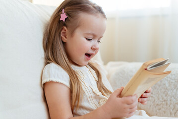 Fototapeta premium Little cute blond girl reading book siting on a sofa. Child reading, dreaming and imagination development. International Children's Book Day. Kids Love to reading. World Book Day