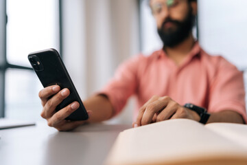 Close-up cropped shot of unrecognizable bearded businessman in pink clothes using phone sitting at office desk with book and work documents by window. Freelancer male texting message on smartphone.