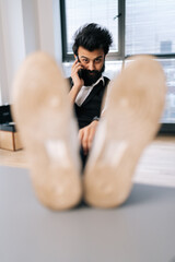 Fototapeta na wymiar Vertical portrait of confident Indian businessman talking on smartphone sitting with feet on table in office by window, looking at camera, sneakers on foreground. Stylish hipster using mobile phone.