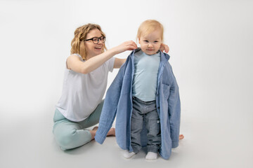 Young funny woman mother putting on oversized denim shirt on little blond blue-eyed girl baby...