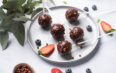Chocolate cake pops with nuts on a plate with fresh berries on a light background. Homemade healthy candies on a stick.