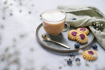 Cappuccino or latte with milk foam in a cup with homemade berry cookies and blueberries on a light background with gypsophila branches. Concept spring morning breakfast.