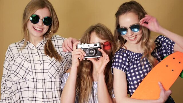 Three beautiful smiling girls with colorful penny skateboards.Women in summer hipster checkered shirt clothes near golden wall.Taking selfie pictures on retro camera. In sunglasses. Slow motion