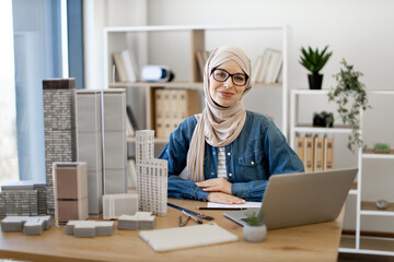 Young muslim woman wearing hijab and spectacles sitting at office desk in real estate agency. Efficient realtor using wireless technologies while arranging purchase of residential properties indoors.