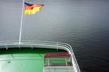 Im Wind wehende deutsche Fahne in Schwarz, Rot und Gold bei Sonnenschein am Heck eines Ausflugsschiff auf dem Edersee bei Waldeck im Landkreis Waldeck-Frankenberg in Hessen
