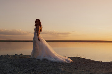 Fototapeta na wymiar Silhouette of bride in white long dress at sunset on sea. Sun horizon. Bride wearing beautiful wedding dress.