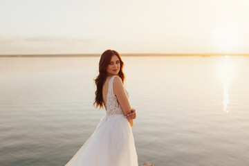 Fototapeta na wymiar Beautiful stylish young bride is posing at the sea view. Wedding ceremony outdoors on the seashore. Summer sunset.
