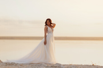 Fototapeta na wymiar Romantic beautiful bride in white dress posing with sea and mountains in background at sunset. Wedding ceremony outdoors on the seashore. 